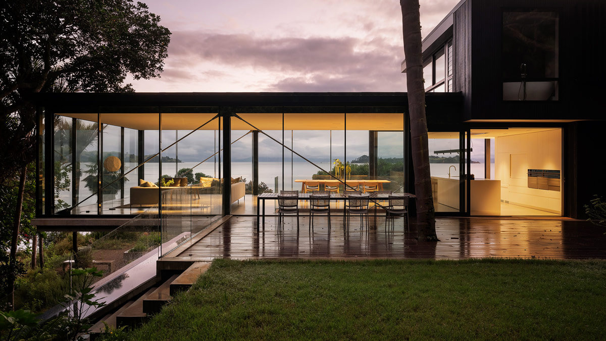 An evening view through windows of Mahuika Beach House lounge, dining, kitchen area to the sea beyond.