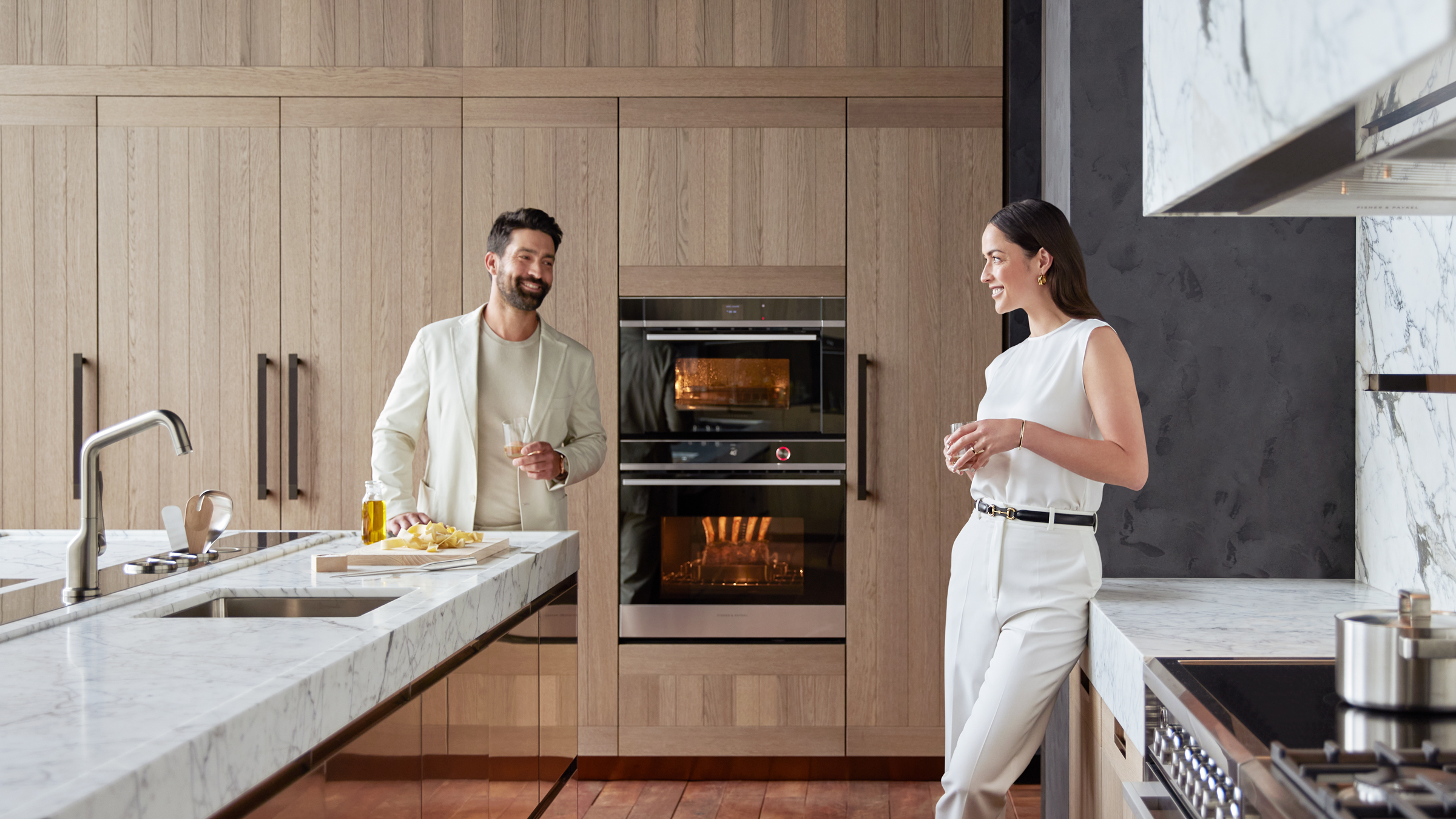 A couple stand conversing in a kitchen featuring light timber cabinetry