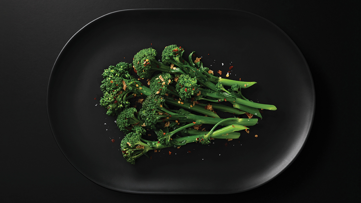Black plate of steamed broccolini garnished with crushed nuts and chilli.