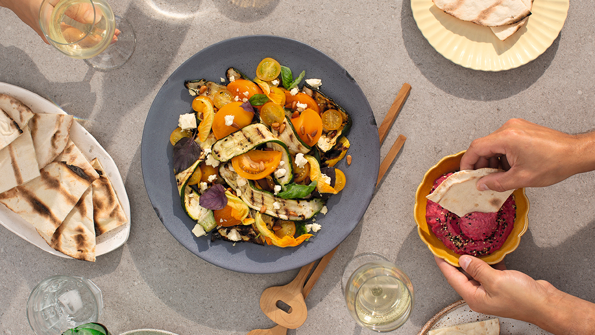 Table set for multiple people to enjoy a charred zucchini salad with flatbread, dip and wine.